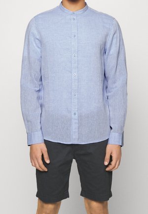 Helles blaues Langarmhemd mit einem Bandkragen, knopflosem Design und weichem, strukturiertem Stoff. Kombiniert mit dunklen Shorts.
