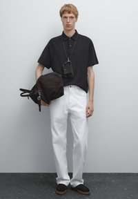 Černé polo tričko s krátkým rukávem, bílé kalhoty rovného střihu, hnědá semišová taška a tmavé boty se světlou podrážkou. Jednoduchý design, minimální akcenty.