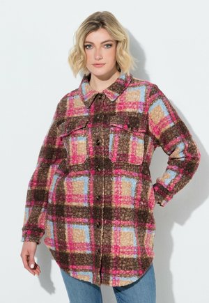 Vrouw met kort blond haar draagt een knoopsluitend, pluizig geruit jasje in roze, bruin, beige en blauw, gecombineerd met blauwe spijkerbroek, poseert met hand op heup.