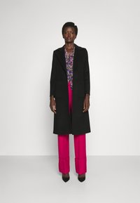 Schwarzer langer Mantel mit Schalkragen, getragen über einer blumigen Bluse und hochtaillierten rosa Hosen. Das Modell steht mit den Händen an den Seiten und trägt Absätze.