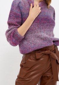 Gestrickter Pullover in Rosa- und Lilatönen mit strukturierendem Muster, hohem Ausschnitt und lockeren, gerippten Bündchen. Braune Hose mit Gürtel.