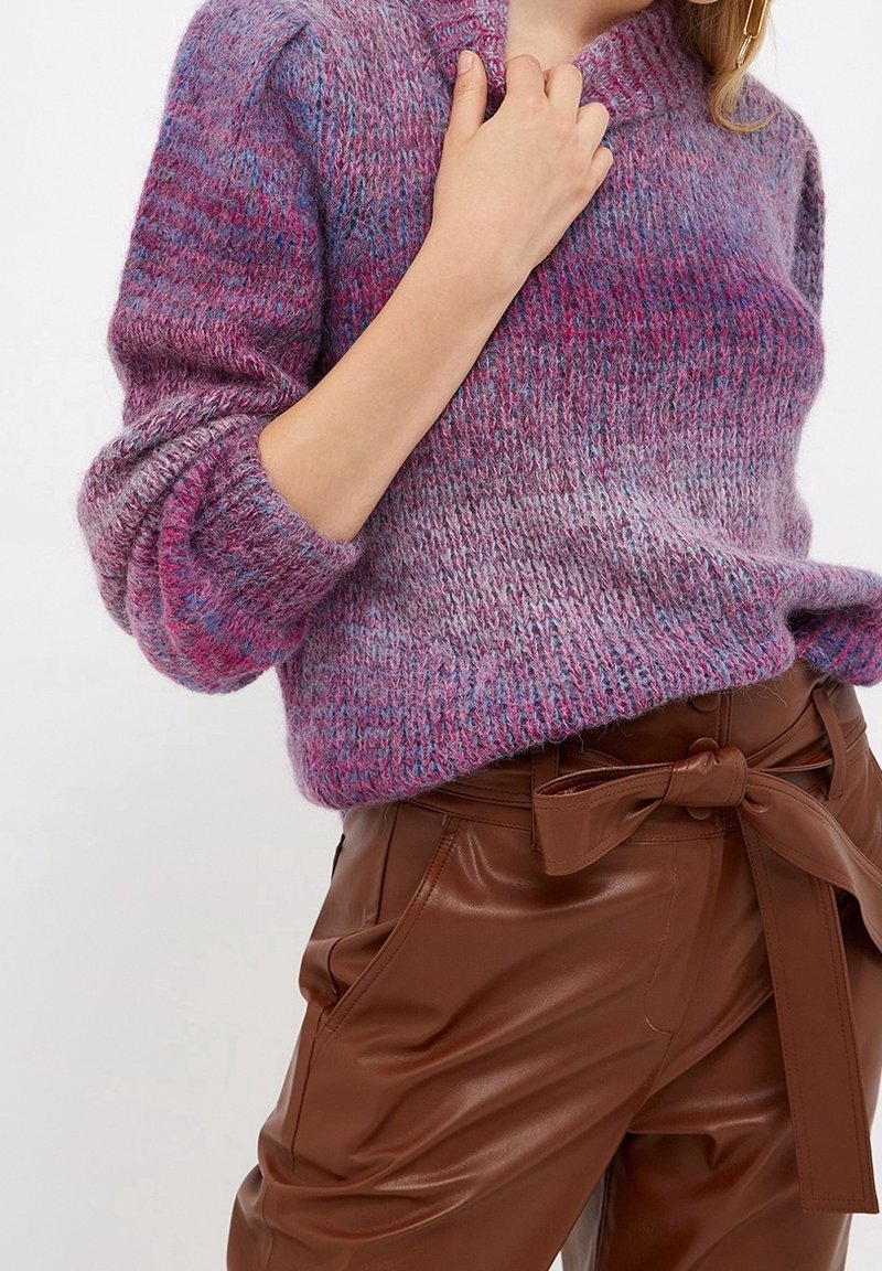 Gestrickter Pullover in Rosa- und Lilatönen mit strukturierendem Muster, hohem Ausschnitt und lockeren, gerippten Bündchen. Braune Hose mit Gürtel.