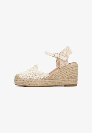 Sandalia de cuña tipo espadrille beige con suela de yute trenzado, puntera tejida y correa ajustable en el tobillo con hebilla dorada y charm.