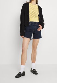 Svart stickad cardigan över en gul spetsstickad topp, kombinerad med mörkblå denimshorts och svarta loafers med kedje-accessoarer. Vita strumpor.