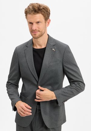 Man in gray textured suit jacket and matching pants fastening the jacket, wearing a black shirt underneath, on a white background.