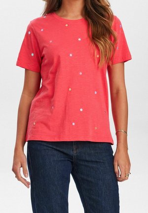 Frau trägt ein korallenfarbenes T-Shirt mit kurzen Ärmeln, verziert mit kleinen, mehrfarbigen Punkten, und dunkelblaue Jeans, dazu Armbänder und Ringe an ihrer rechten Hand.