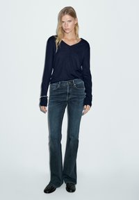 Vrouw die tegen een witte muur staat, gekleed in een zwarte langermouw V-hals top, donkere flared jeans en zwarte schoenen met een zilveren armband.