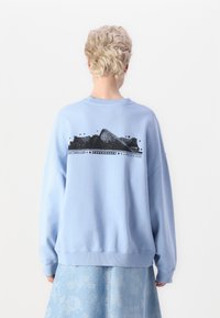 Lys blå genser med rund hals, oversized passform og teksturert grafisk trykk på baksiden med abstrakte former og tekst.