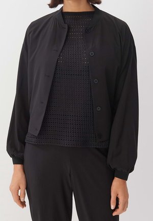 Femme portant une veste noire boutonnée sur un haut en résille noir à manches bouffantes et un pantalon noir assorti, debout devant un fond uni.