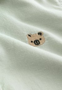 Tessuto verde chiaro con superficie testurizzata che presenta una piccola faccia di orso ricamata in toni beige e neri.