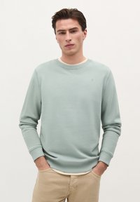 Lysegrøn sweatshirt i blødt stof med rund hals og ribbede manchetter. Modellen har den på over en hvid skjorte, hænderne i lommerne, neutral baggrund.