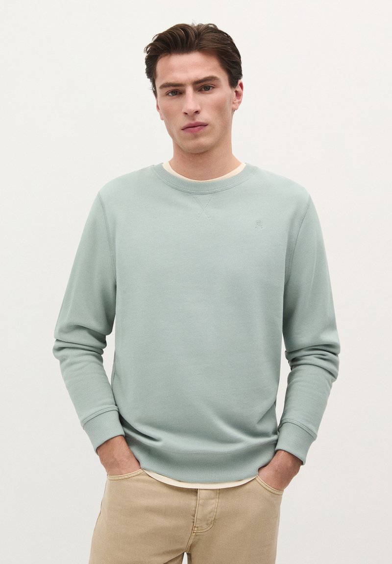 Lysegrøn sweatshirt i blødt stof med rund hals og ribbede manchetter. Modellen har den på over en hvid skjorte, hænderne i lommerne, neutral baggrund.