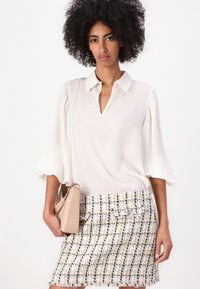 Blusa blanca con mangas plisadas, combinada con una falda de tweed multicolor con detalles de botones. La modelo sostiene un bolso beige.