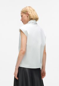 Blonde woman wearing sleeveless white top with studded high collar and black leather skirt, facing left against white background.