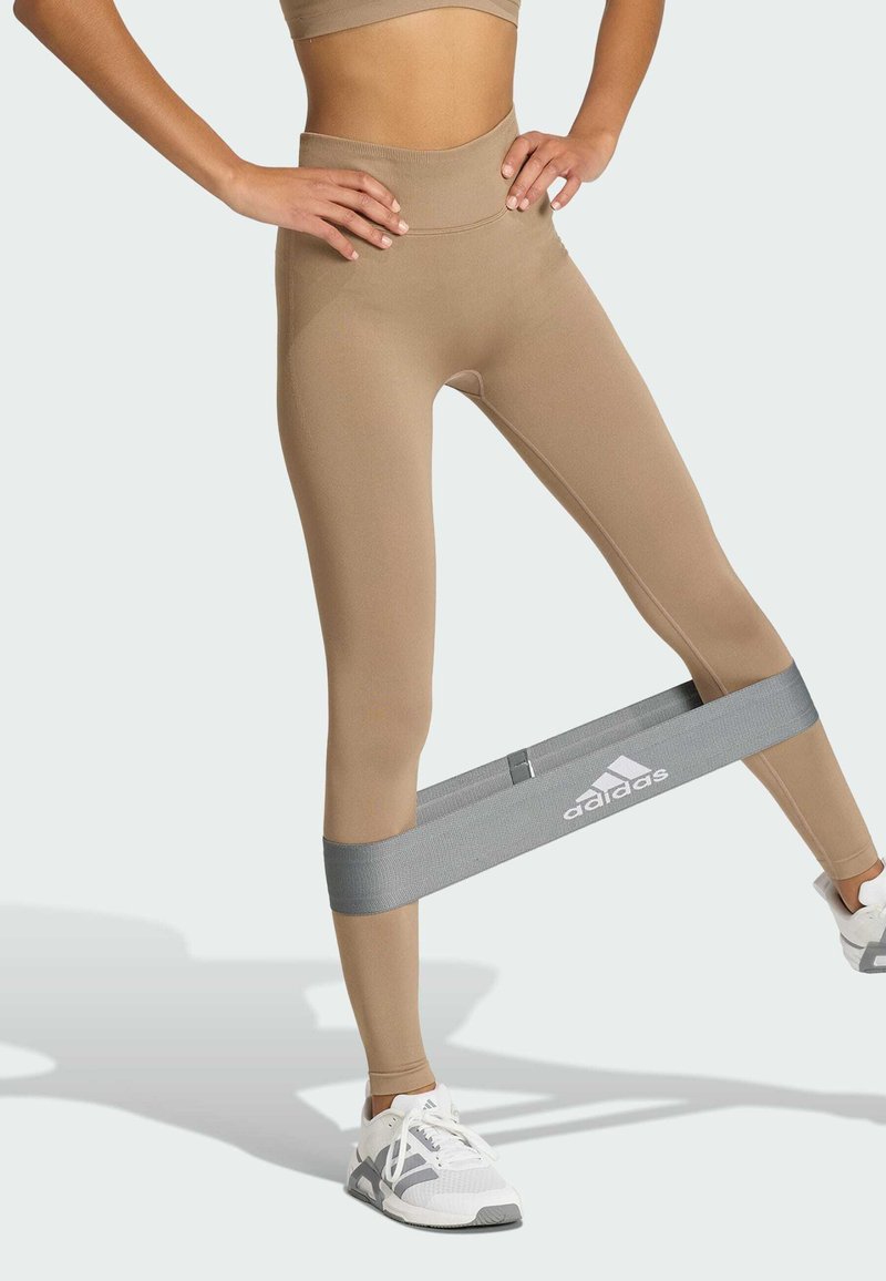 Partie inférieure du corps d'une personne en leggings beige et baskets blanches en train de faire de l'exercice avec une bande de résistance grise Adidas autour des jambes.