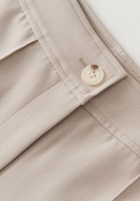 Tessuto beige chiaro con una texture liscia, dotato di una tasca cucita e di un bottone rotondo color crema con due fori vicino al bordo.