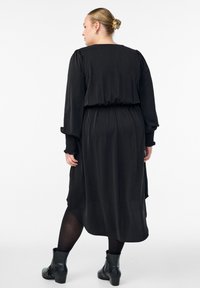 Mujer con cabello rubio recogido en un moño, vistiendo un vestido largo negro, medias negras y botas negras de tobillo, de pie con la espalda hacia adelante.