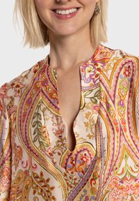 Blouse présentant un motif cachemire floral pastel, tissu doux, encolure fendue, et manches courtes avec des détails de design complexes en rose, vert et orange.