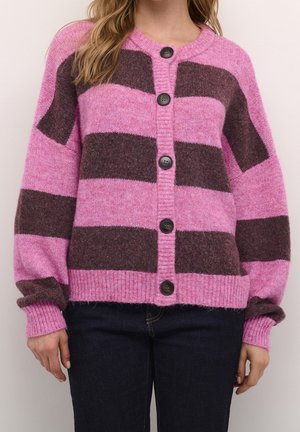Femme portant un cardigan tricoté rayé rose et marron avec des boutons noirs, associé à un jean bleu foncé, debout devant un fond uni.