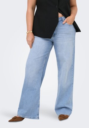 Person som har på seg lyseblå vide jeans, brune spisse sko, svart ermeløs topp, med en hånd i lommen og gullarmbånd på håndleddet.