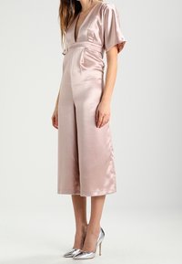 Bledě růžový saténový overal s hlubokým V-výstřihem, krátkými rukávy, opaskem v pase a širokými nohavicemi. Spárováno s metalickými stříbrnými vysokými podpatky.