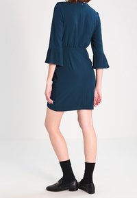 Teal klänning ovanför knäet med veckade klockärmar och en figursydd midja. Slät tyg med en hög ryggkrage och detaljer i sidsömmen.