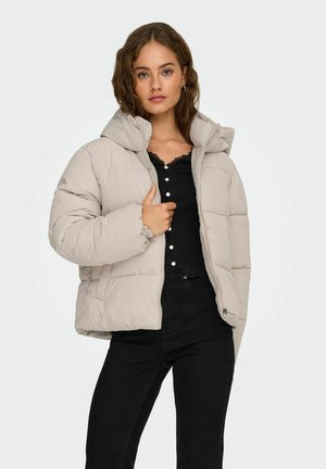 Beige puffer jacket with a hood, featuring a quilted design and front zipper, paired with a black top and dark jeans.