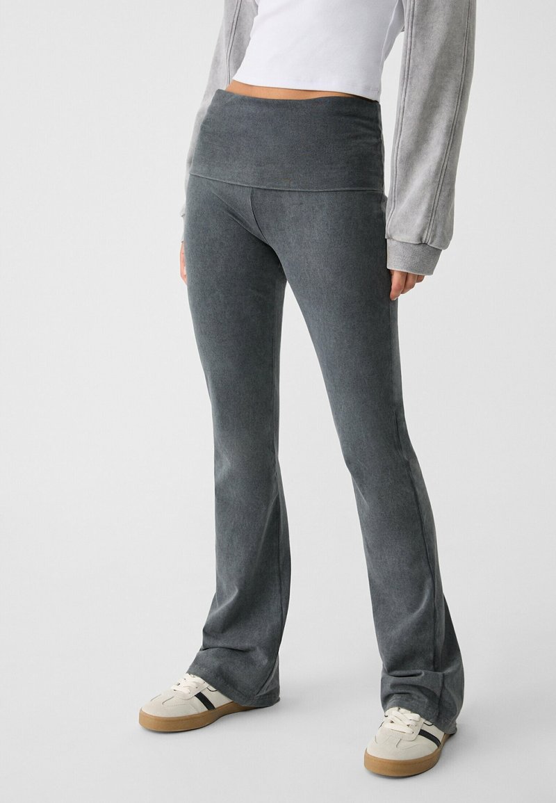 Personne portant un pantalon évasé taille haute gris foncé, un haut court blanc, une veste grise à manches longues et des baskets blanches avec des rayures noires.