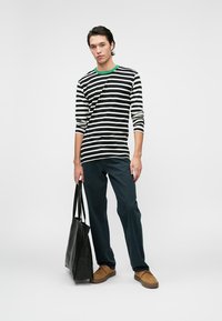 Svart-hvitt stripete langermet skjorte med grønn krage, paret med mørkeblå bukser og brune sko; holder en svart tote bag.