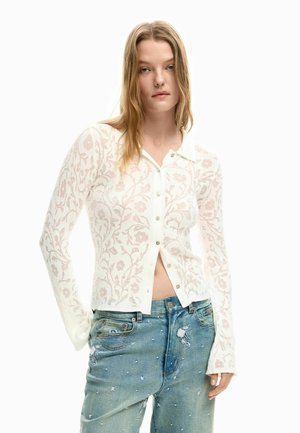 Biała przezroczysta kardigan na guziki z wzorem kwiatowym i kołnierzykiem, połączona z jasnoniebieskimi zniszczonymi dżinsami z ozdobami i delikatnym strzępieniem.