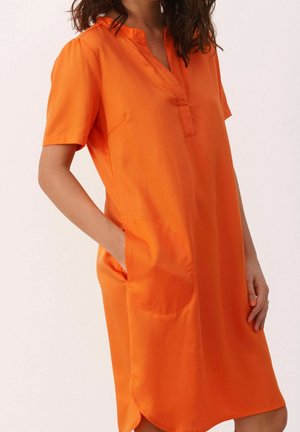 Femme portant une robe orange à manches courtes, longueur genou, avec une fente au col, une main dans la poche latérale, sur un fond clair uni.