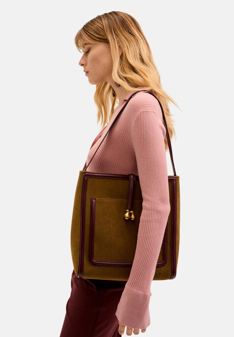 Sac à corduroy moutarde avec une bordure bordeaux, doté d'une poche avant et de détails en métal doré, porté par un mannequin de profil.