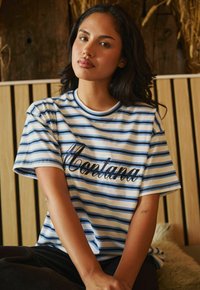 Camiseta de algodón a rayas con líneas horizontales azules y blancas; presenta "Montana" en texto cursivo negro en el pecho; corte relajado.