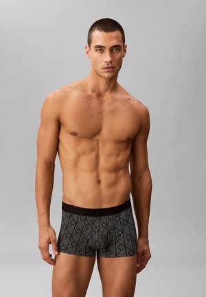 Uomo muscoloso in piedi davanti a uno sfondo grigio, indossa boxer aderenti con motivo bianco e nero.