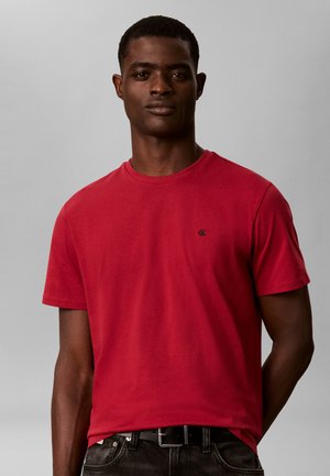 Mężczyzna w prostym czerwonym t-shircie z krótkim rękawem i małym czarnym logo na piersi, czarnym pasku i ciemnych spodniach na szarym tle.