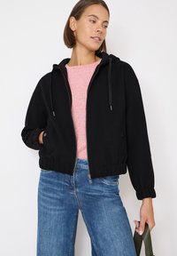 Sudadera negra corta con cremallera frontal y cordones ajustables, combinada con un suéter rosa y jeans de pierna ancha azules. Textura suave.