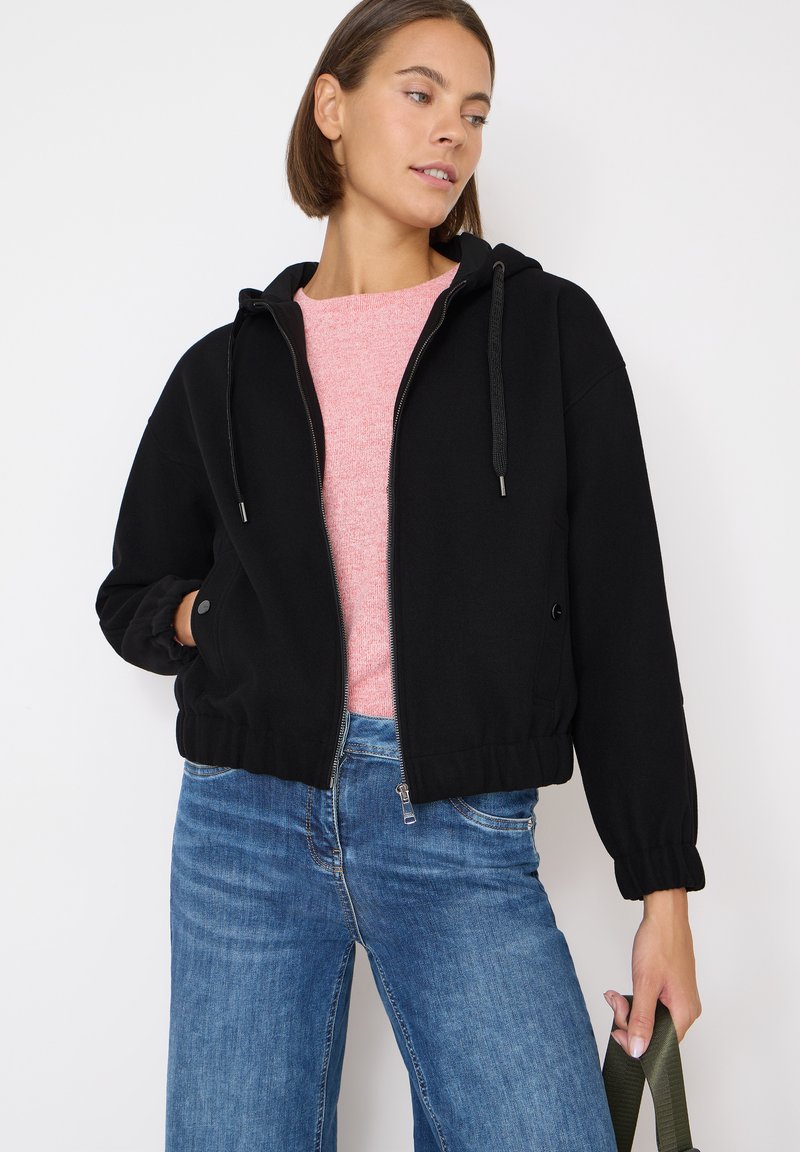 Sudadera negra corta con cremallera frontal y cordones ajustables, combinada con un suéter rosa y jeans de pierna ancha azules. Textura suave.