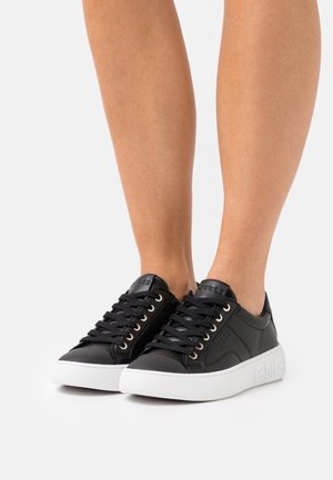 Paire de baskets noires basses avec semelles blanches épaisses portées sur jambes nues, avec des lacets noirs et un logo discret de la marque sur les côtés.
