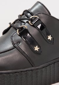 Chaussure en cuir noir avec une semelle plateforme texturée, dotée d'un design à lacets, d'accents noirs brillants et d'un détail en matériel doré en forme d'étoile.