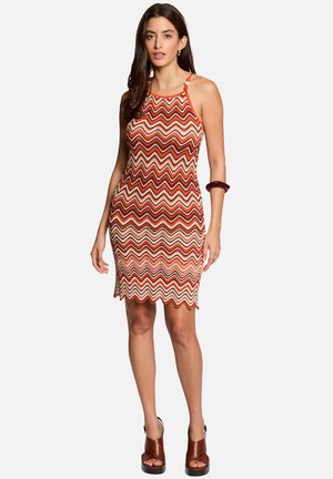 Kvinde iført ærmeløs knælængde kjole med røde, orange og hvide bølgede striber, brune højhælede sandaler og mørkt armbånd på venstre arm.