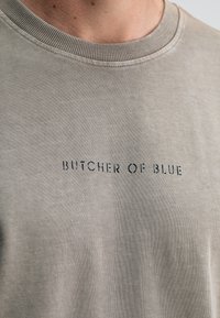 Grau Baumwoll-T-Shirt mit Rundhalsausschnitt und feiner Textur, mit dem Aufdruck "BUTCHER OF BLUE" auf der Vorderseite.