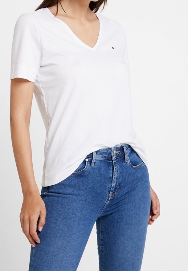 Camiseta blanca de cuello en V de algodón, de mangas cortas, con un pequeño logo en el pecho, combinada con jeans de denim azul.