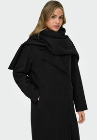 Cappotto nero in lana con colletto oversize e dettaglio a sciarpa. Caratterizzato da una linea dritta, maniche lunghe e tasche laterali. Tessuto morbido.