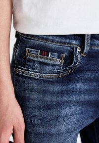 Dark blue denim jeans with a classic fit, featuring a small pocket with a striped detail in red and white. Distressed accents are present.