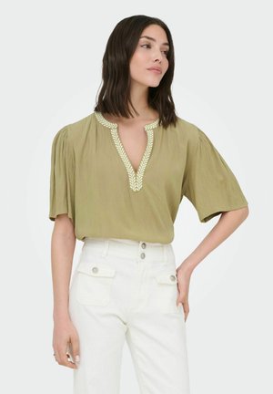 Femme aux cheveux foncés mi-longs, portant un chemisier vert olive clair à décolleté en V avec bordure de feuilles et un pantalon blanc taille haute avec poches boutonnées.