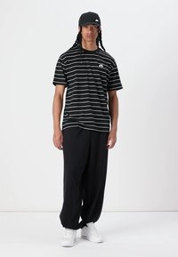 Schwarz-weiß gestreiftes T-Shirt mit kleinem Logo, kombiniert mit lockeren schwarzen Hosen und weißen Sneakers. Das Modell trägt eine schwarze Mütze.