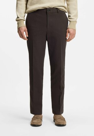 Mann trägt schwarze Anzughose, beigefarbenen Pullover und beige Slipper, steht vor weißem Hintergrund.