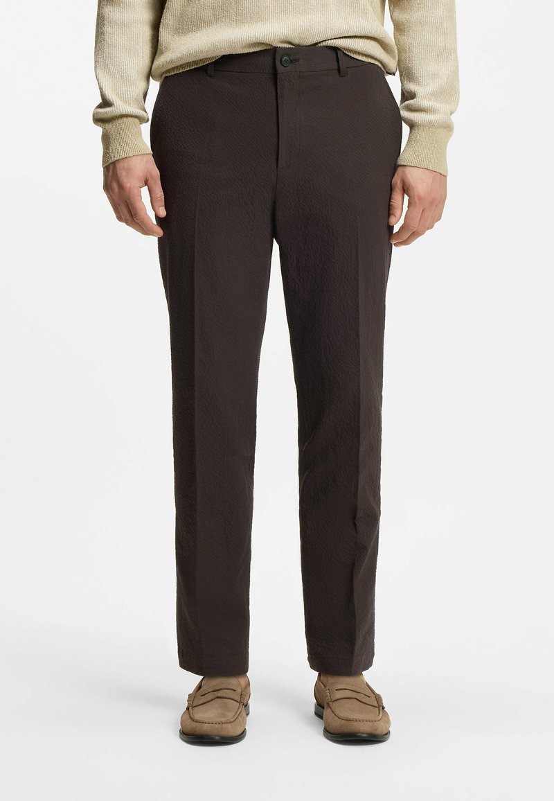 Mand iført sorte bukser, beige sweater og lysebrune loafers, stående foran en hvid baggrund.