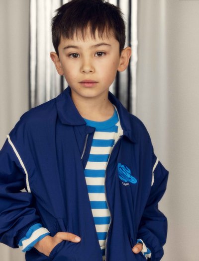 Νεαρός αγόρι με κοντά σκούρα μαλλιά, φοράει μπλε μπουφάν πάνω από μπλε και λευκή ριγέ μπλούζα, στέκεται με τα χέρια στις τσέπες του μπουφάν.