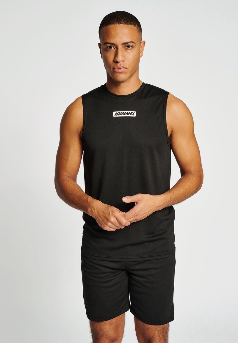 Homme debout en maillot de sport noir sans manches et short avec le logo « HUMMEL » sur le devant du maillot, mains jointes au niveau de la taille.